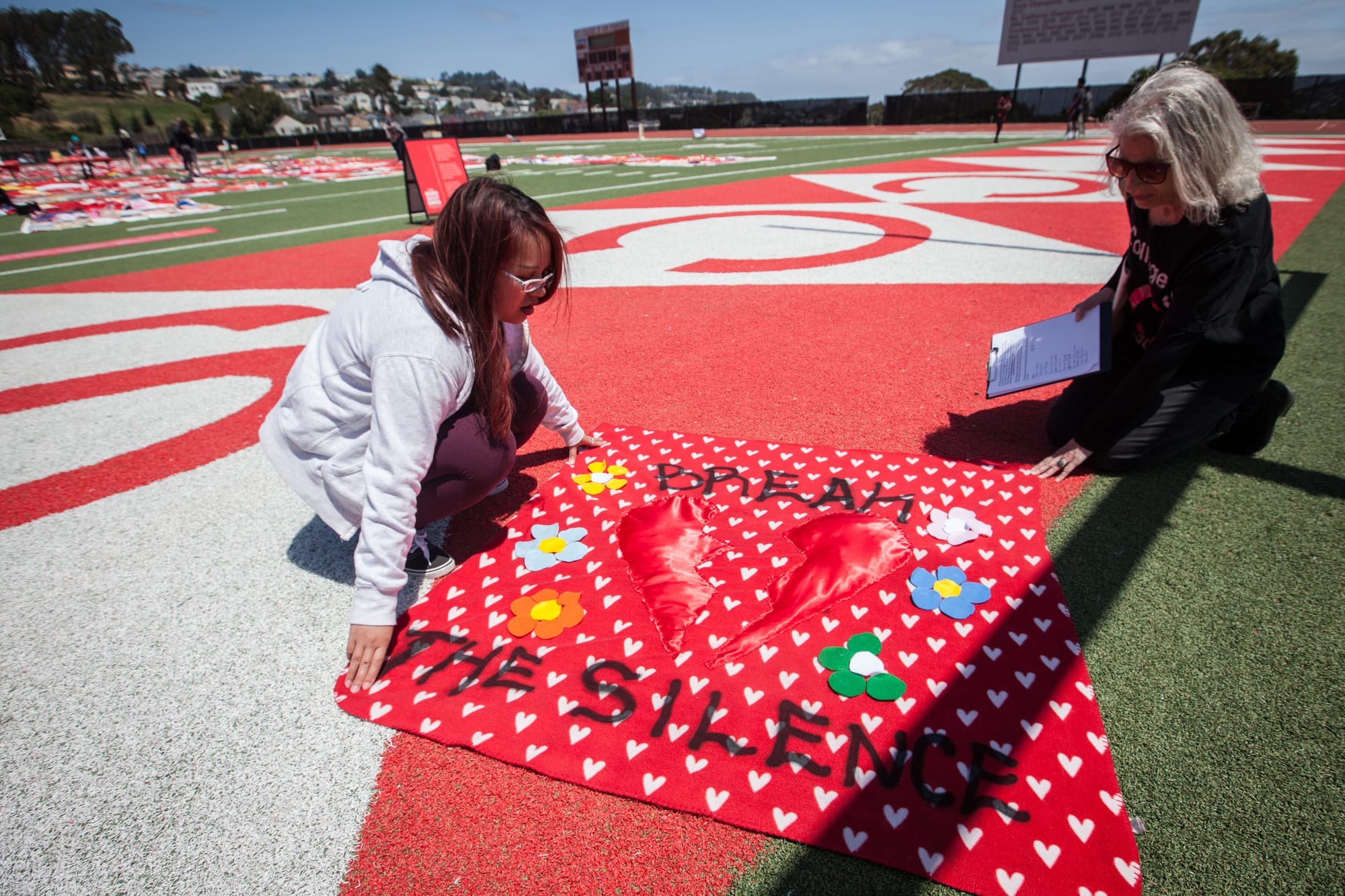 (L-R) An instructor of Ending Sexual Violence: Peer Education Shella Cervantes and Interim Project SURVIVE Coordinator Leslie Simon display a red square quilt made by a CCSF student Valentina Vargas during an installation of The Monument Quilt on the football field of George M. Rush Stadium at City College of San Francisco Ocean Campus on Saturday, May 6, 2017. Vargas made the quilt out of her old blanket that she had since she was 16 years old and figured that the blanket had seen all her past unhealthy relationship; so making it into a quilt is the way for her to recycle old energy through healing and art. (Photo by Ekevara Kitpowsong) 